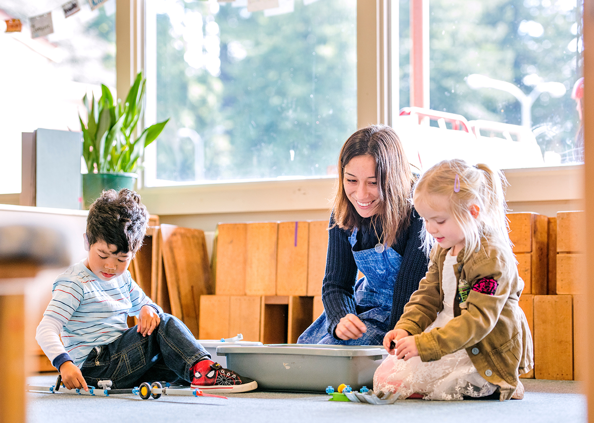 A woman engaging with two children on the ground as they play with their toys.